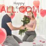 blue color T-shirt Man offering red rose bouquet to his girlfriend on bed with room decorated with red color heart shaped balloons.